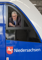 Thomas Nawrocki (im Anzug), Bereichsleiter Fahrzeugmanagement bei der LNVG schaut schmunzeln und stolz aus dem seitlichen Zugführerfenster des neuen Fahrzeuges