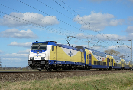 Blau-weiß-gelber Zug der metronom nach links fahrend auf freier Strecke mit blauem Himmel und einzelnen Wolken